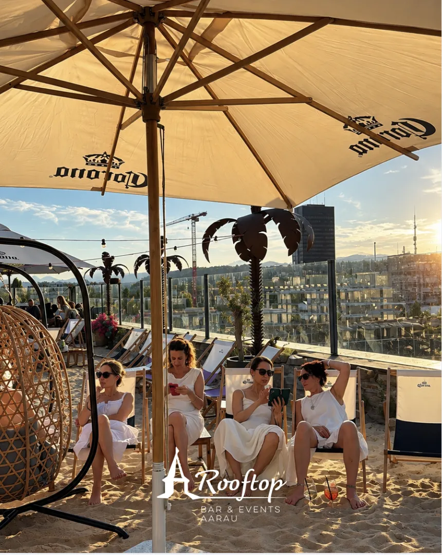 Gäste in Liegestühlen unter dem Sonnenschirm in der Sandoase des Rooftop Aarau bei Abendsonne